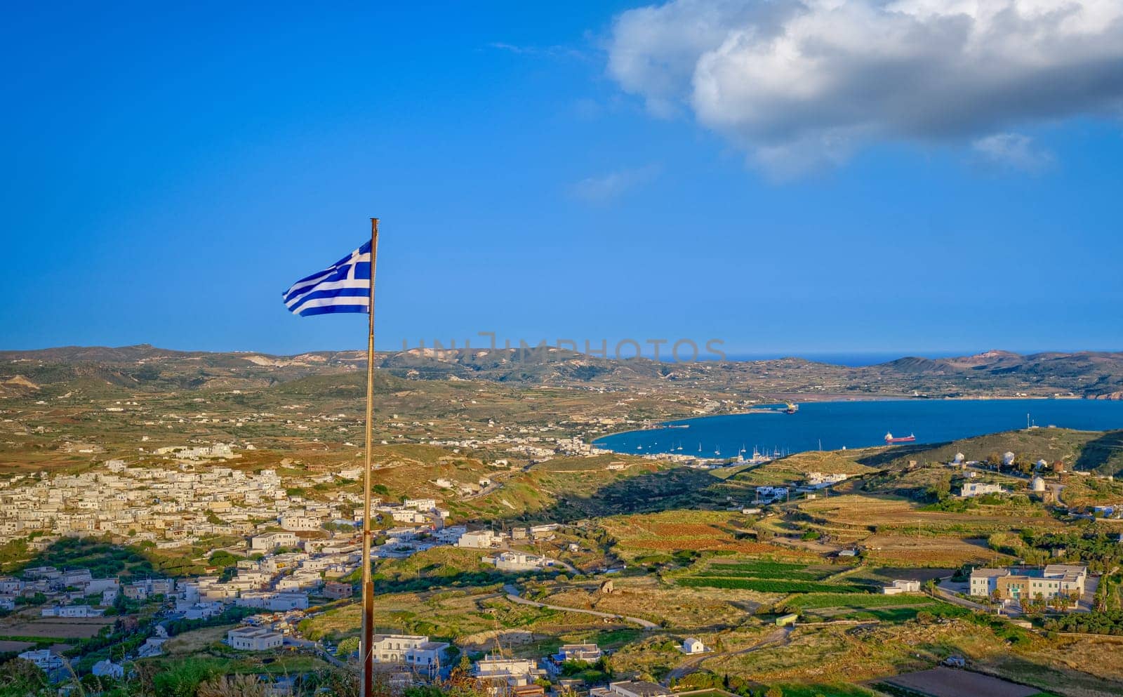 National flag of Greece waving over landscape of Milos island. Adamantas town, harbour and villages at sunset. Famous whitewashed houses, green hills and blue sea, distant mountains, clear sky with cloud, summer sunshine. Mediterranean vacations