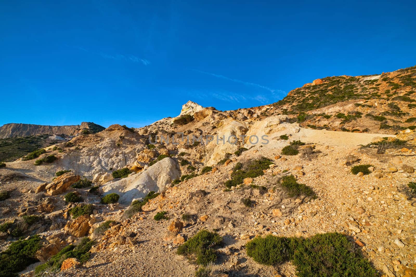 Unique natural rock formations of yellow and orange cliffs in low sunlight, Milos, Greece. Weather-shaped slopes of rocks on Agios Ioannis beach at clear blue sky.