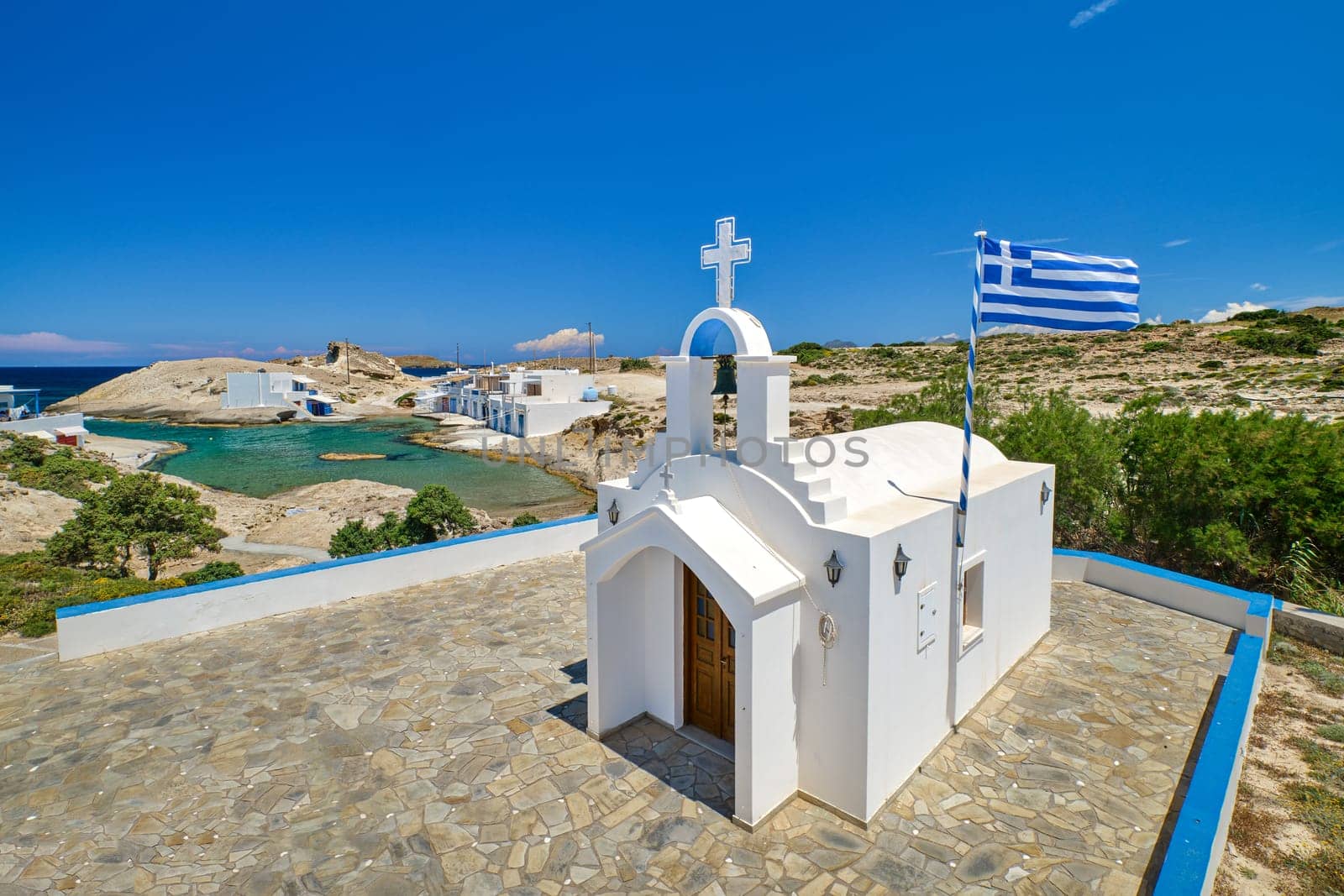 White Greek Orthodox chapel or church and Greek national flag on top of hill on sunny summer day. Typical Greek island, summer landscape, traditional religious building, aerial view, azure bay waters, clear blue sky, few houses by water, Milos Greece