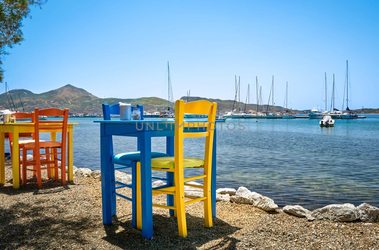 Colorful chairs and tables of Greek tavern by waterfront at sunny day. Vacations in Greece, summer, restaurant, outdoor dining, port, marina, yachts and boats, Mediterranean relax, Cyclades and Aegean islands. Selective focus, blurred background.