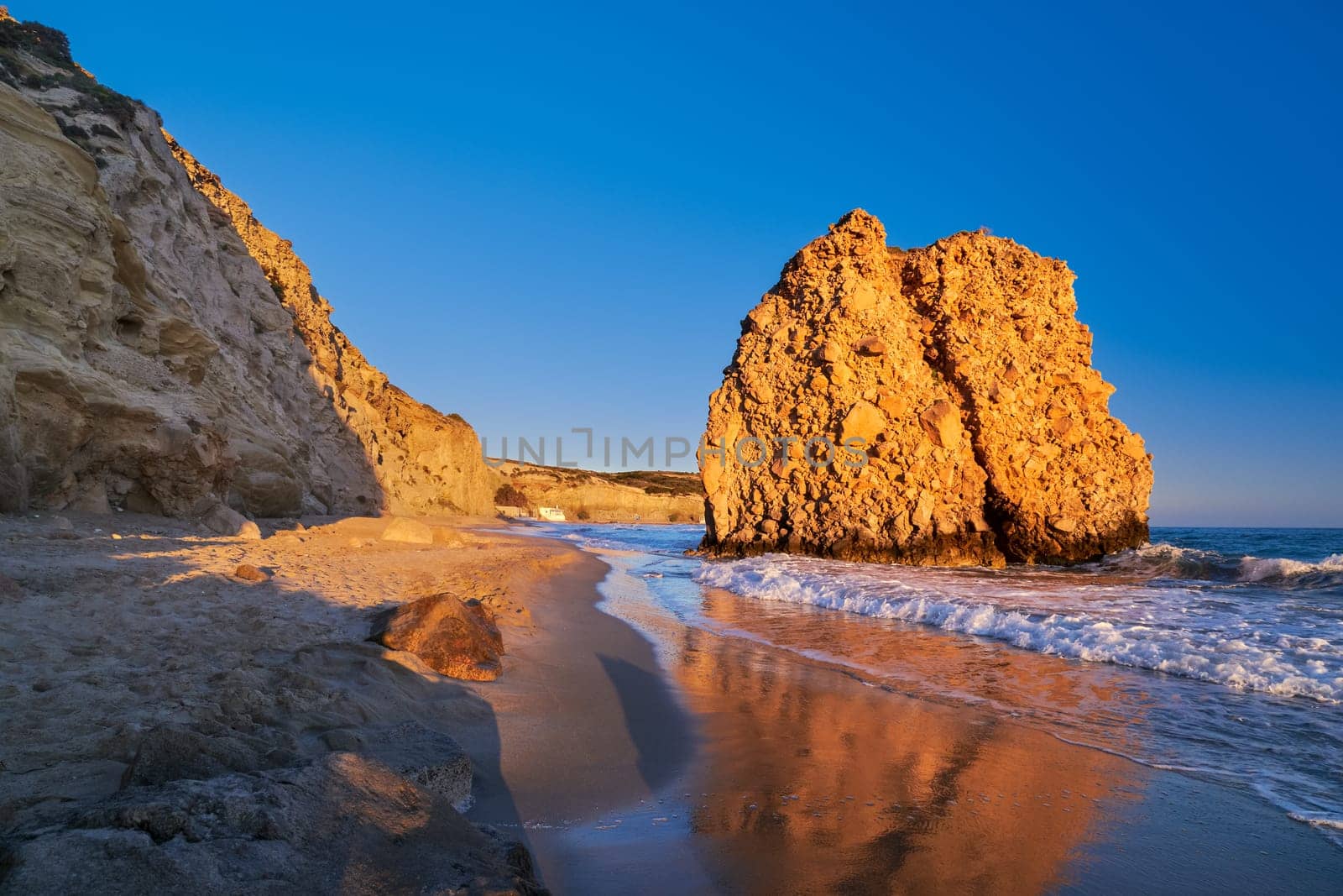 Colorful sunset over Fyriplaka beach, Milos island, Cyclades, Greece. Unique lonely rock on sandy beach lit by low sun mirroring in wet sand of tide. Beautiful sky, rocky hills, sea waves spray, no people.