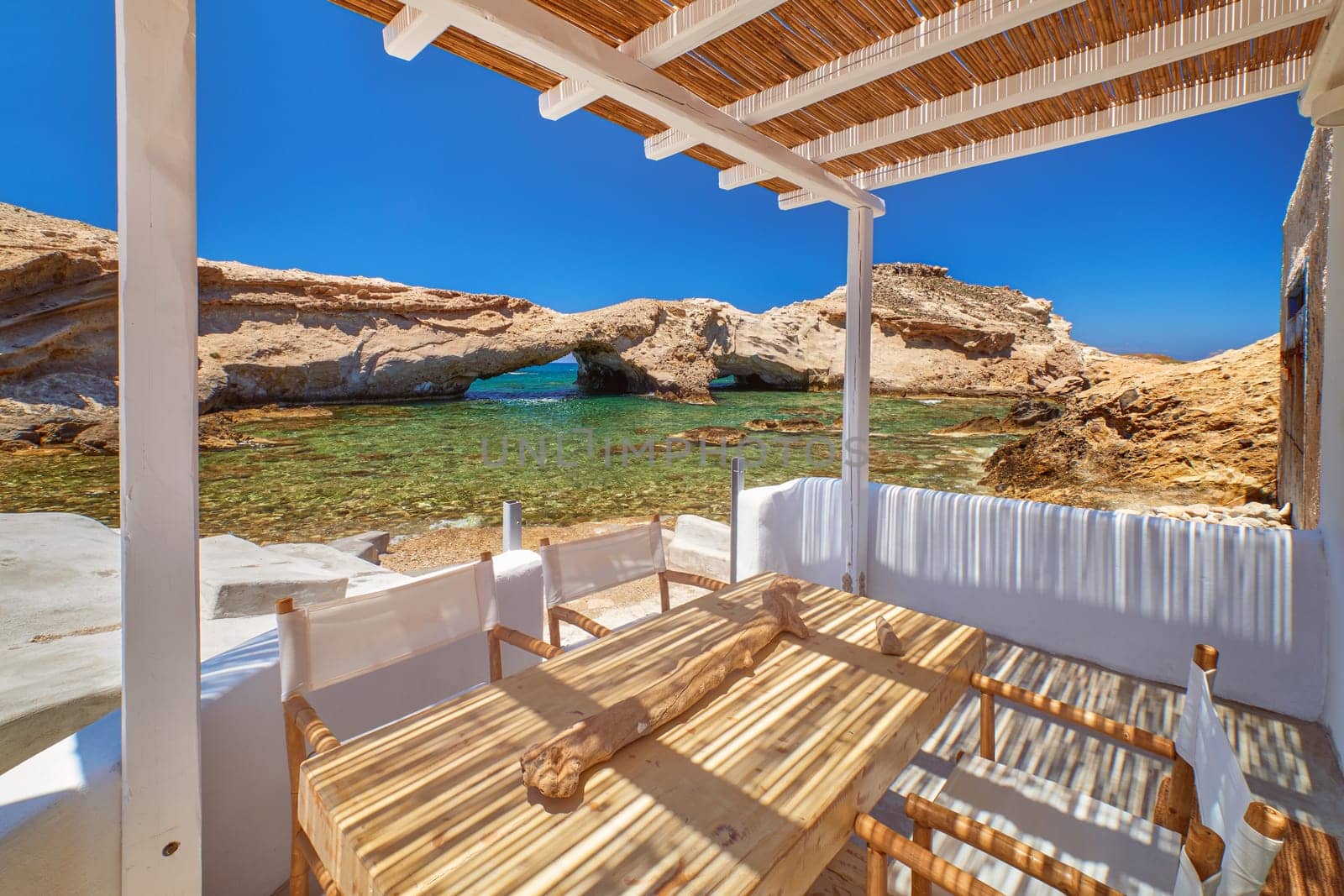 Summer villa by seafront on sunny day. Small private bay, no people, azure clear sea waters, clear blue sky, sunshine, rocks and caves. Terrace, shade, minimalistic design, eco trend, relax, meditation, retreat, Mediterranean lifestyle. Milos, Greece