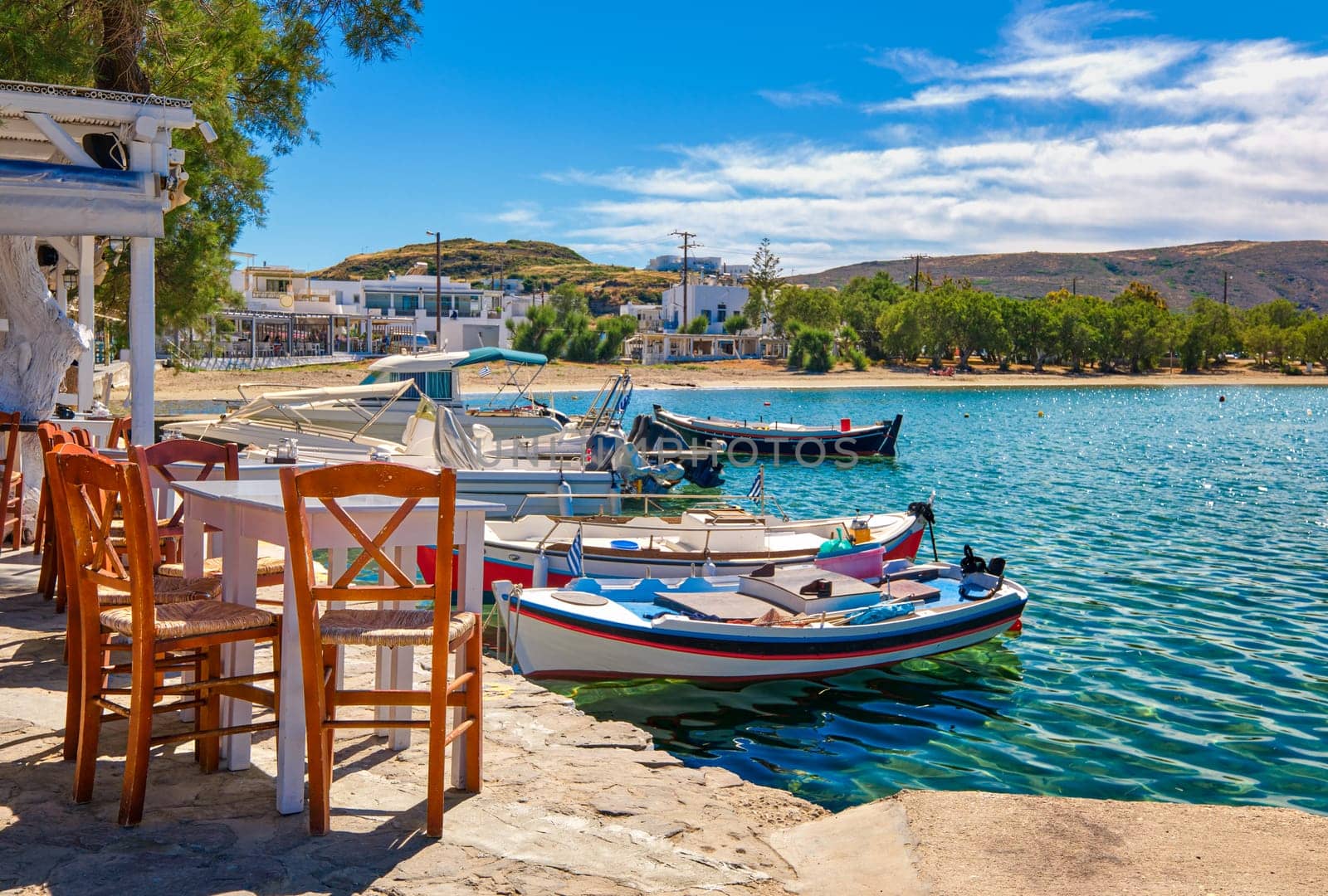Colorful chairs and tables of Greek tavern by waterfront at sunny day. Vacations in Greece, summer, restaurant, outdoor dining, port, marina, yachts and boats, Mediterranean relax, Cyclades and Aegean islands. Selective focus, blurred background.