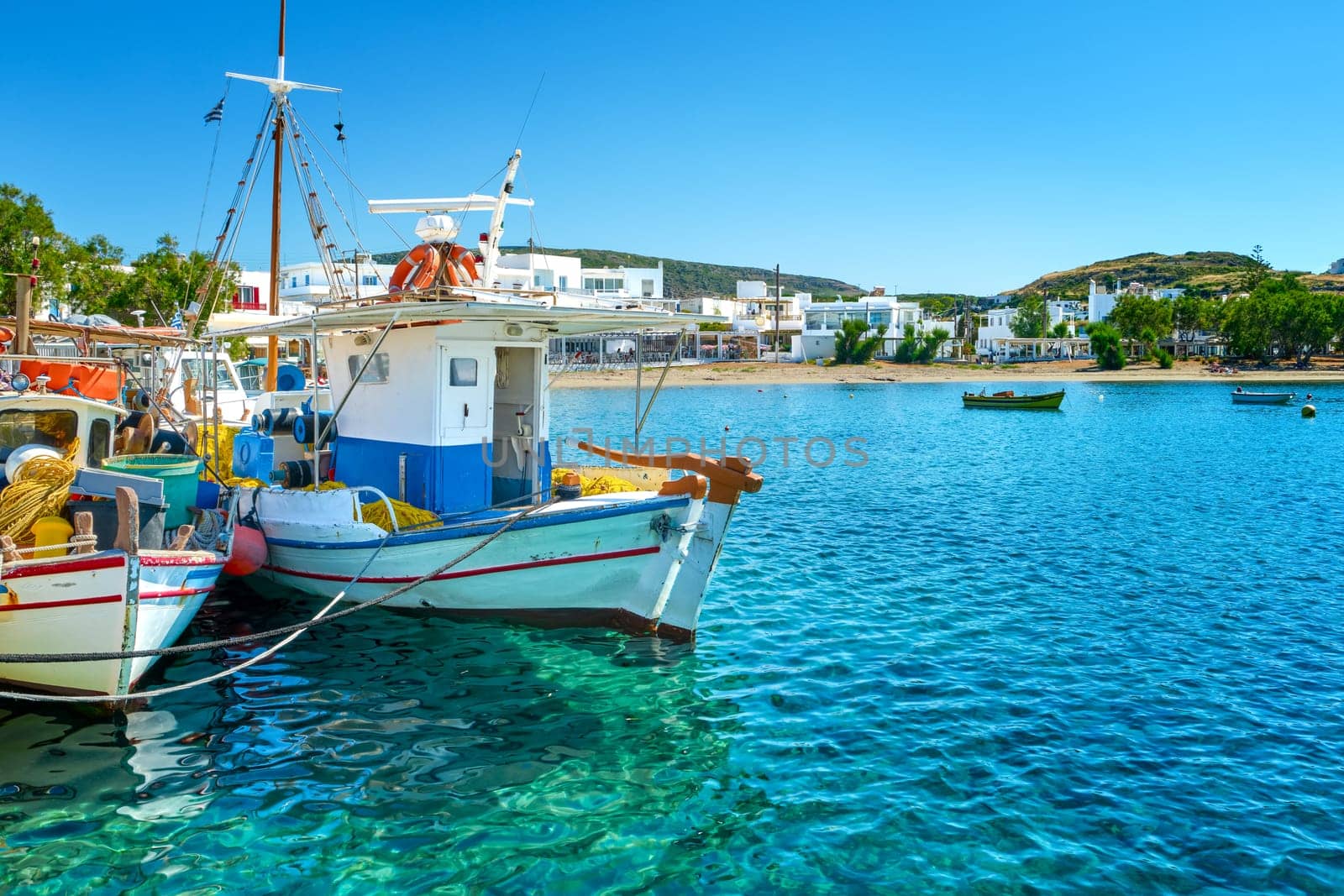 Beautiful view of Greek fishing boats anchored in a small village. Summer day sunshine, island, village, town, whitewashed houses, colorful fishing boats, Mediterranean lifestyle, green hills, vacations, relax, travel destinations. Milos, Greece.