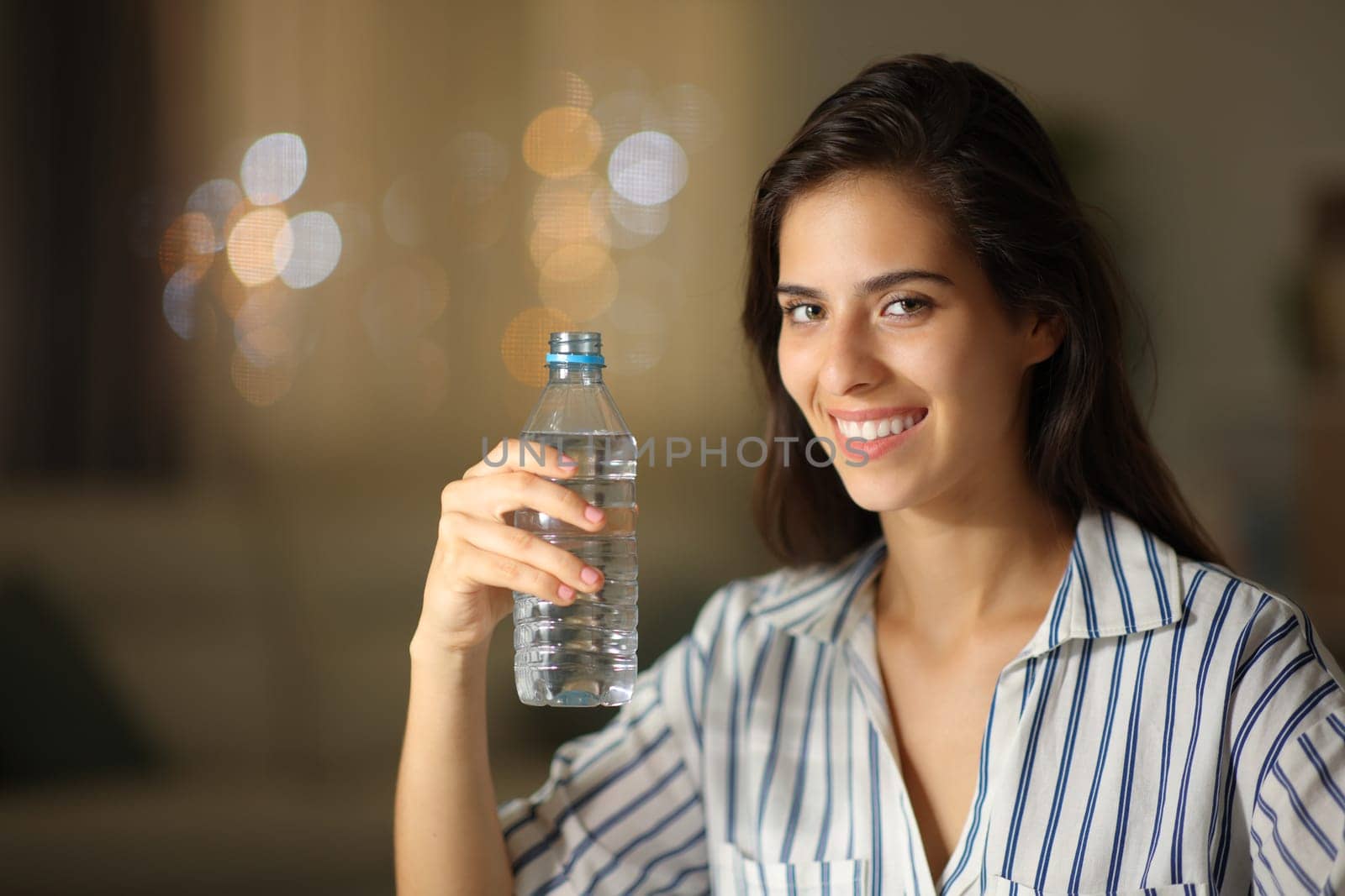 Happy woman posing holding water bottle looking at camera at home in the night