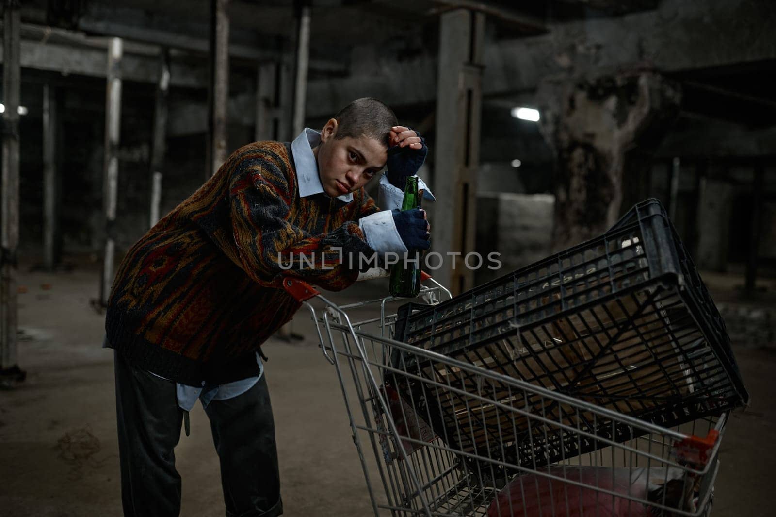Portrait of unhappy young woman immigrant or beggar drinking beer leaned on trolley cart with her belongings