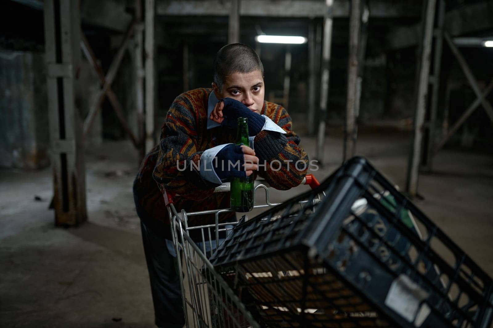 Portrait of unhappy young woman immigrant or beggar drinking beer leaned on trolley cart with her belongings