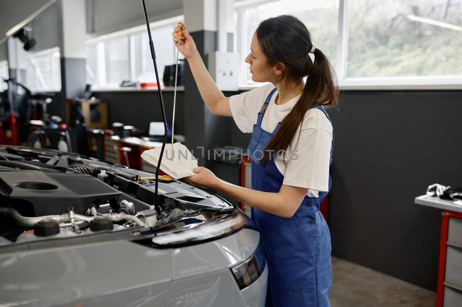 Young woman technician checking oil level in engine using dipstick in car mechanical workshop
