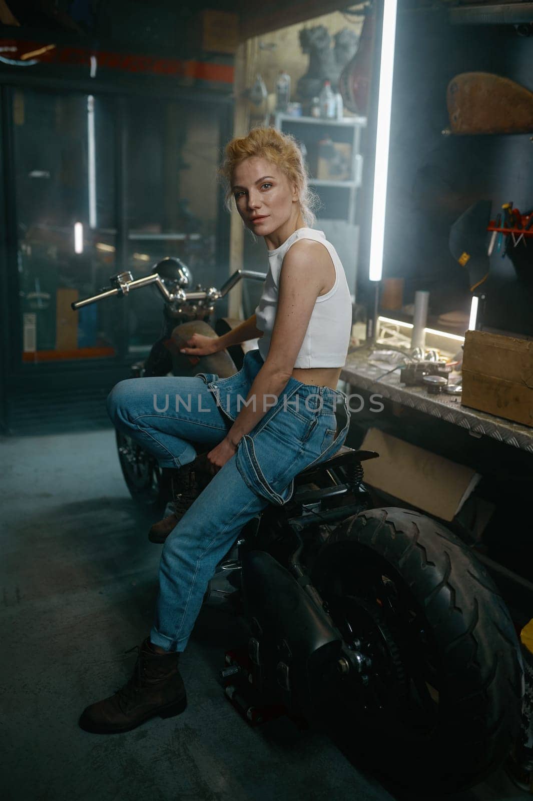 Portrait of beautiful woman biker sitting on motorcycle in garage. Pretty female mechanic posing for camera at workshop