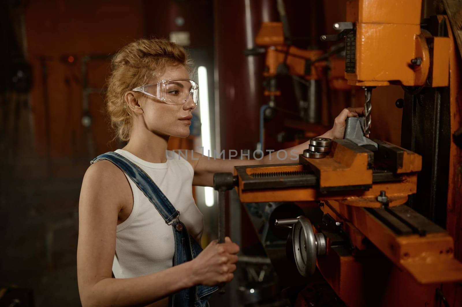 Close up view of focused professional serious young woman wearing safety protective goggles working on electric milling and drilling machine at garage workshop