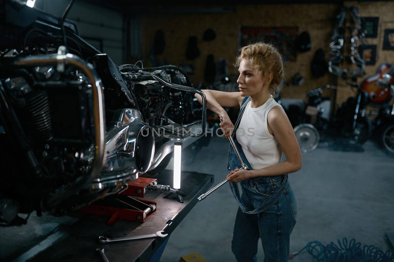Talented girl wearing blue jumpsuit using work tool to repair motorcycle in garage. Pensive young woman at creative authentic workshop