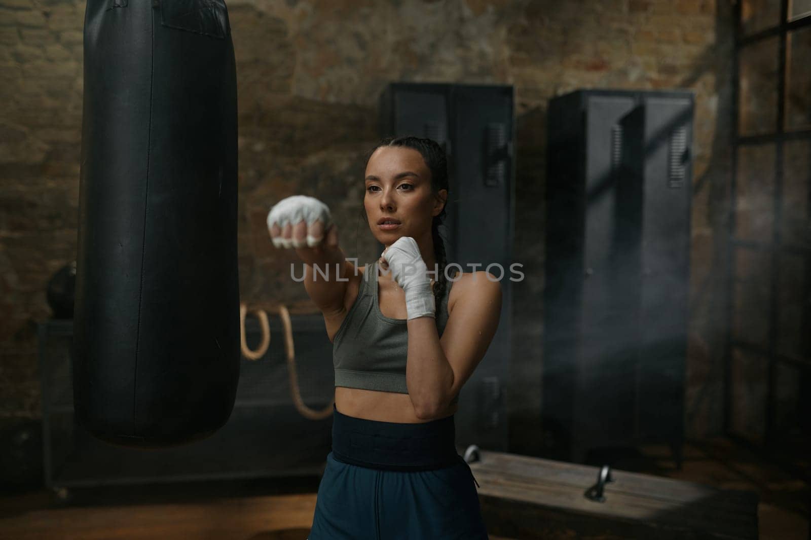 Front view portrait of powerful serious woman boxer in boxing bandage fighting to camera making direct hit. Young fit female fighter training punches workouting at gym