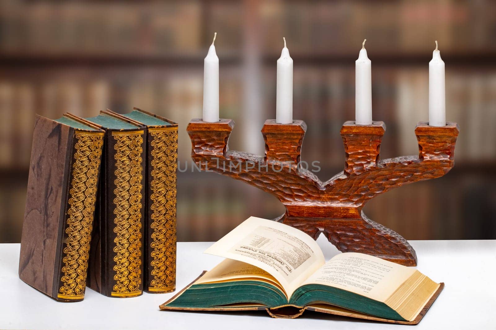 Antique books with an open book in front of an antique wooden candlestick on a bright table over abstract blurred library. University, history, memory and knowledge concept.