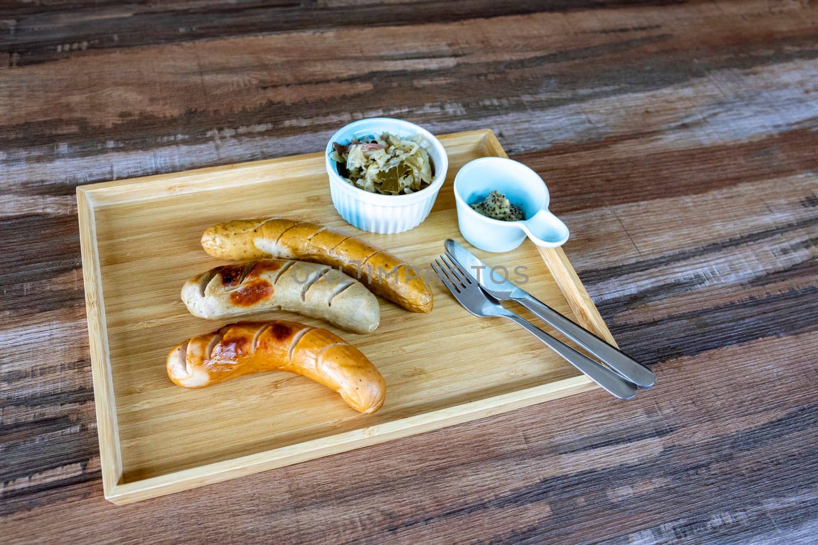 Photos of Grilled German Sausages with sauerkraut and dijon mustard in a small bowl with knife and fork Together in a rectangular wooden tray, on a wood grain table, there is a copy space on the right.