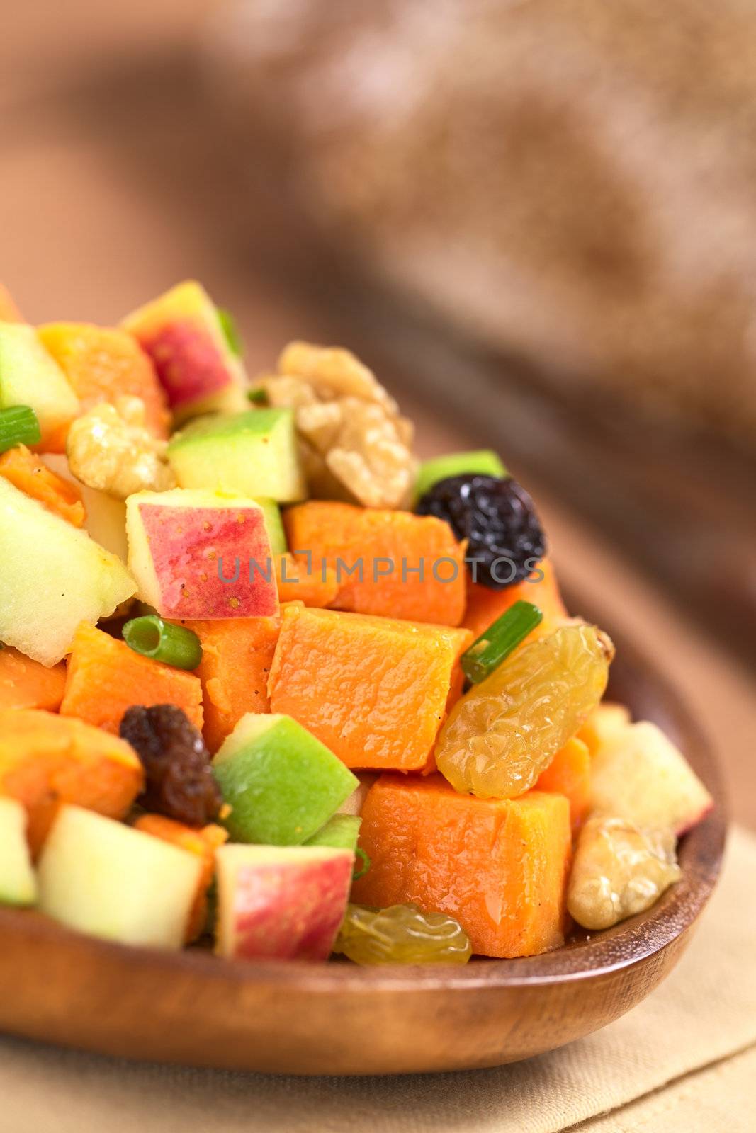 Fresh salad made of cooked sweet potatoes, fresh apples, nuts, raisins and shallots on wooden plate with wholewheat bread in the back (Selective Focus, Focus one third into the salad)