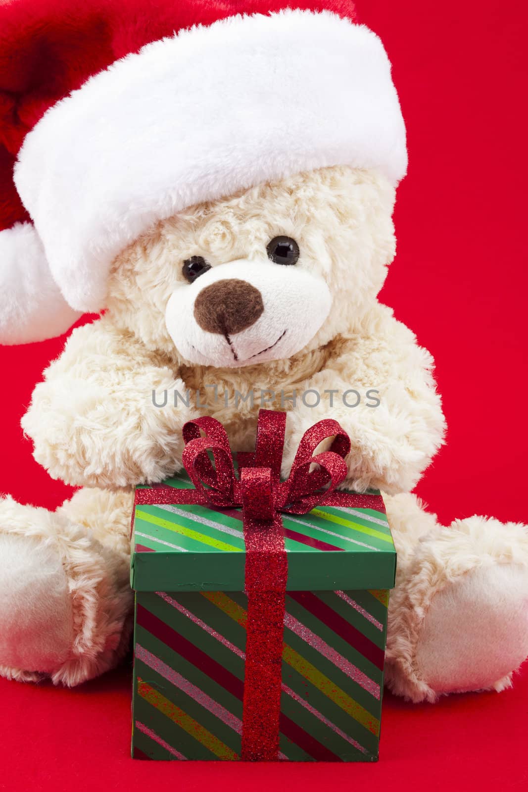 Teddy bear wearing a Santa hat is sitting in front of a unopened gift, on a red background. 