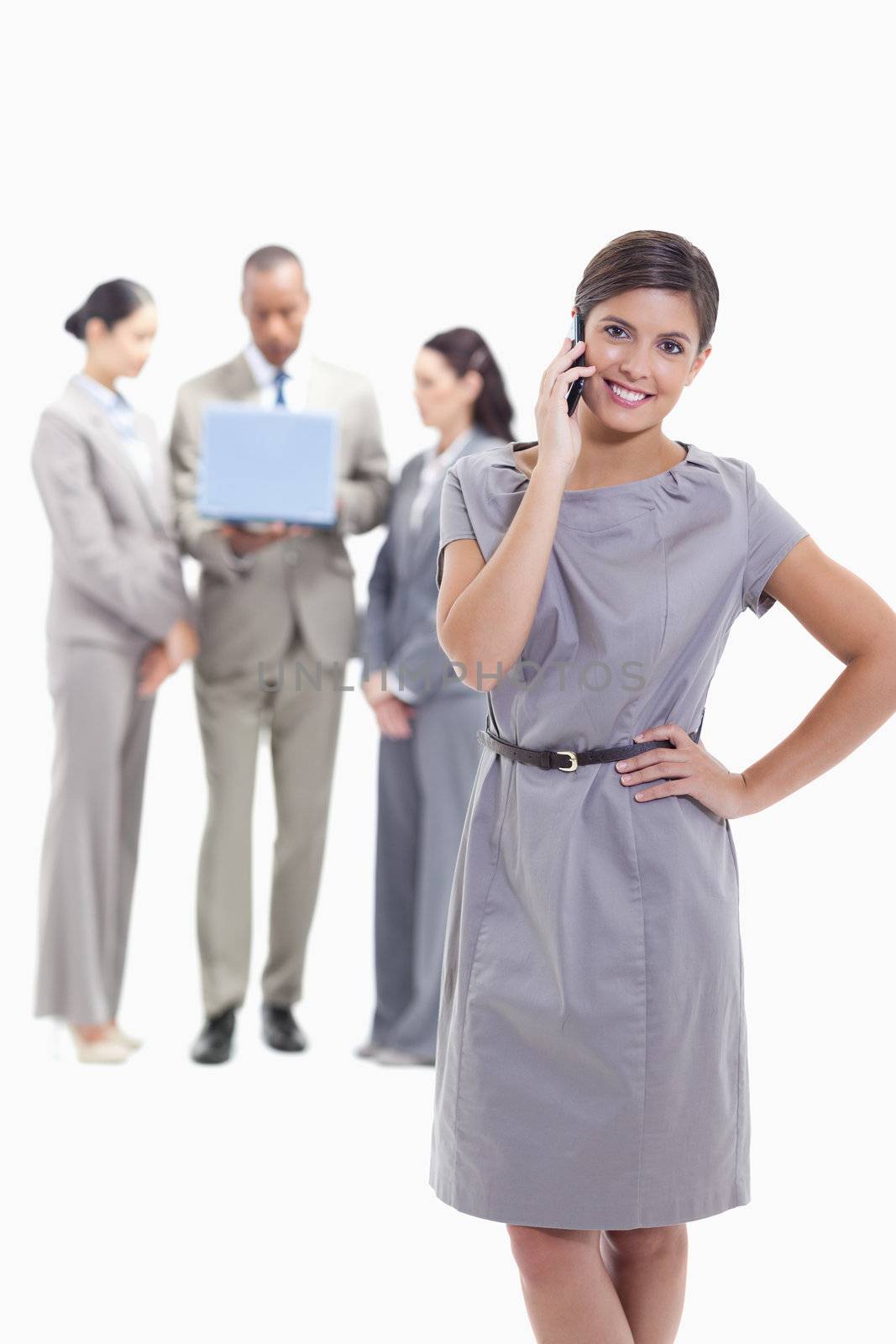 Businesswoman on the phone with a hand on her hip, her legs crossing and her head tilted with co-workers in the background watching a laptop screen