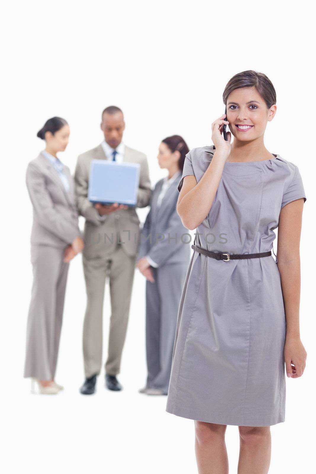 Businesswoman smiling on the phone and looking straight ahead with one arm along her body and co-workers in the background