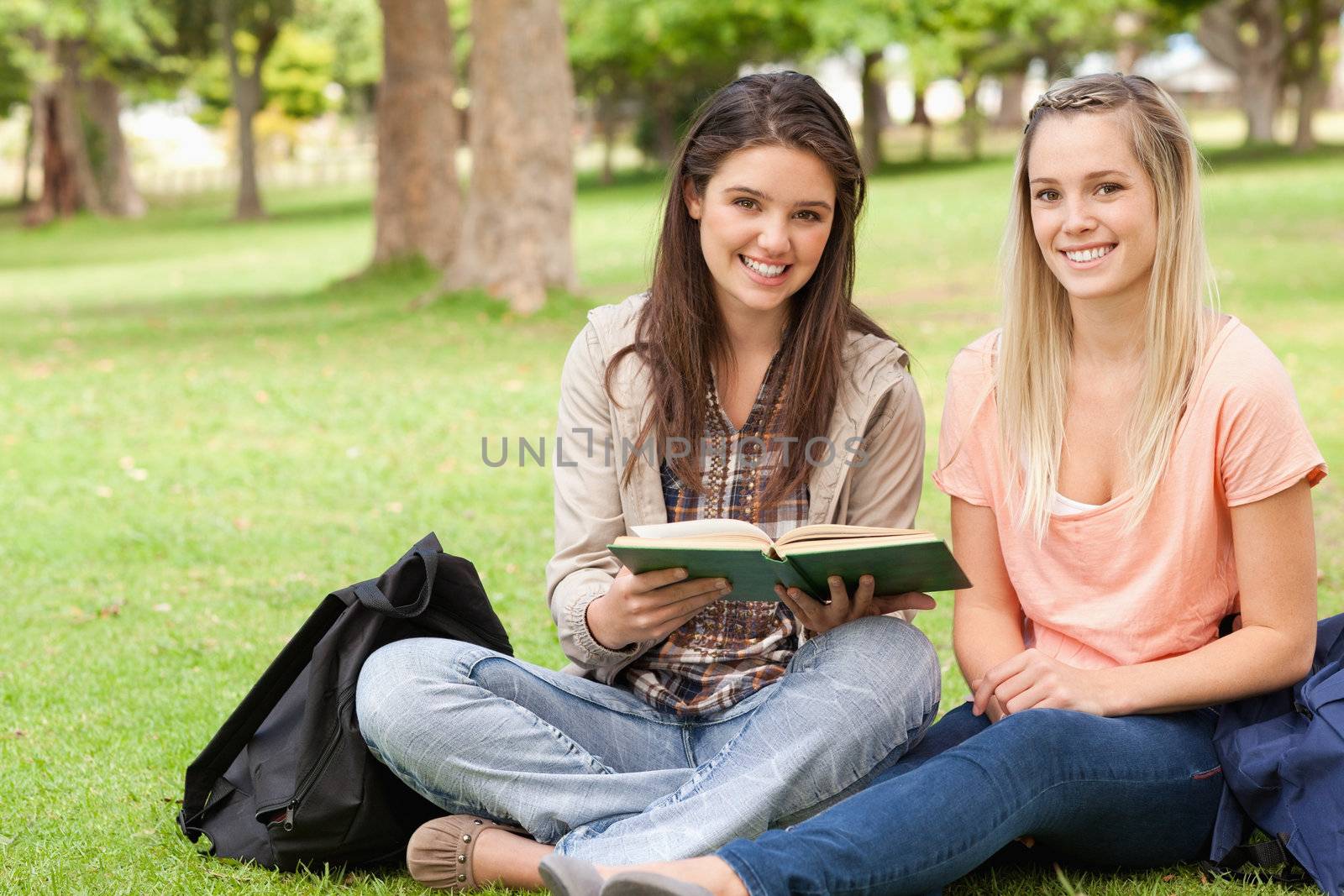 Female teenagers sitting with a textbook in a park while looking at camera