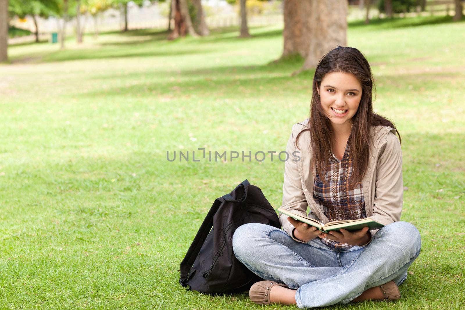 Smiling teenager sitting while holding a textbook in a park
