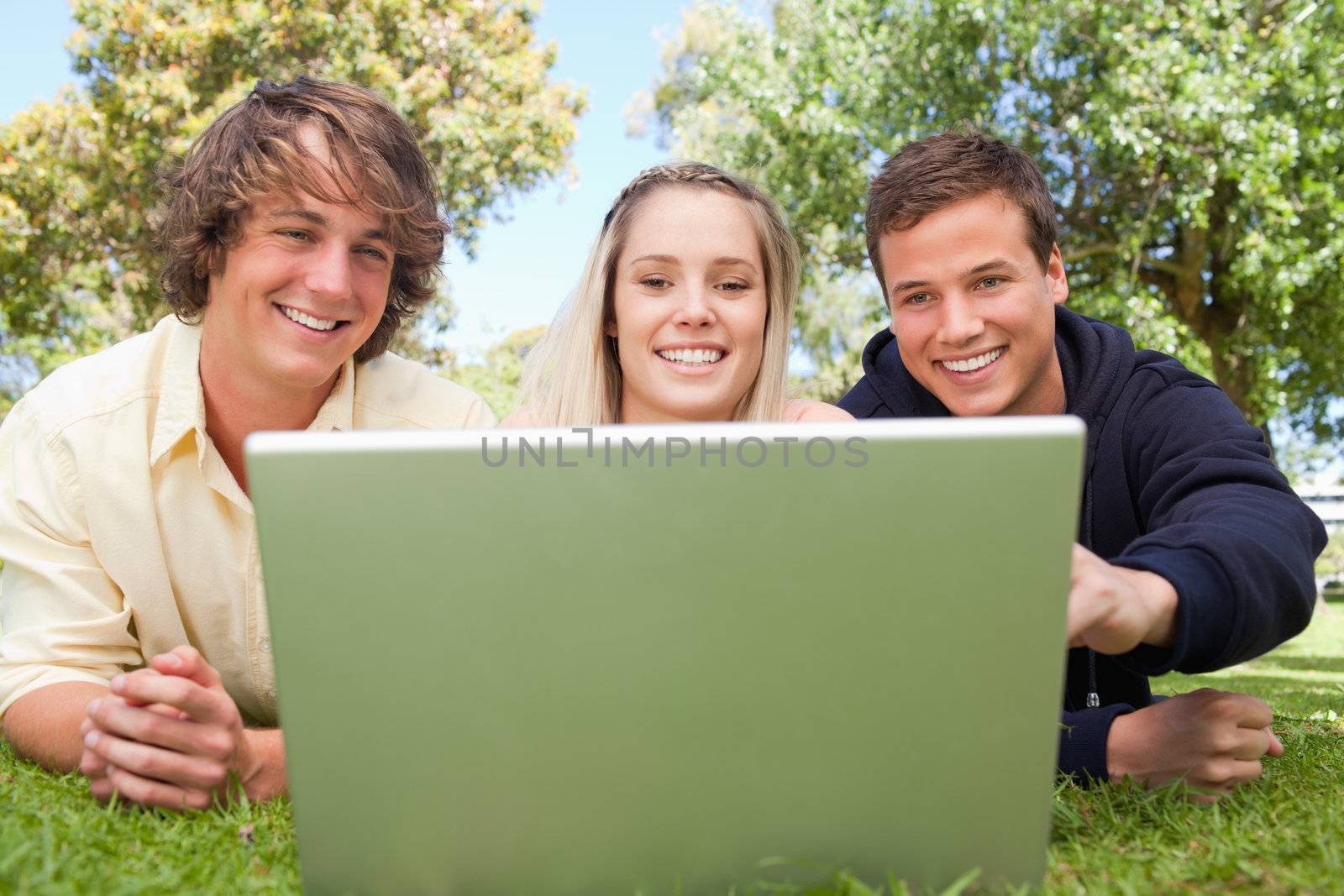 Portrait of three happy students in a park lying while using a laptop