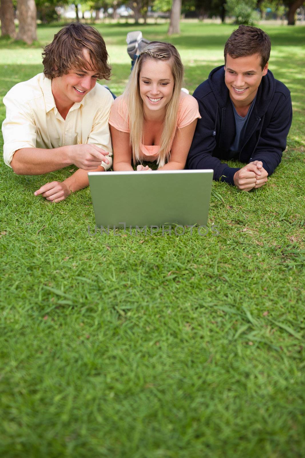 Three happy students in a park lying while using a laptop