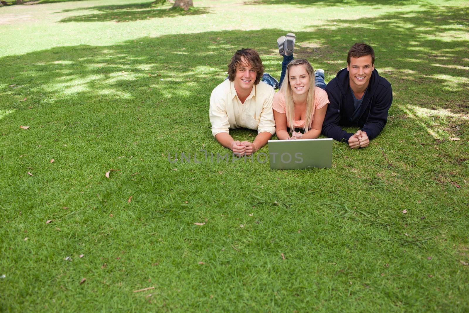 Three smiling students looking at camera in a park lying while using a laptop
