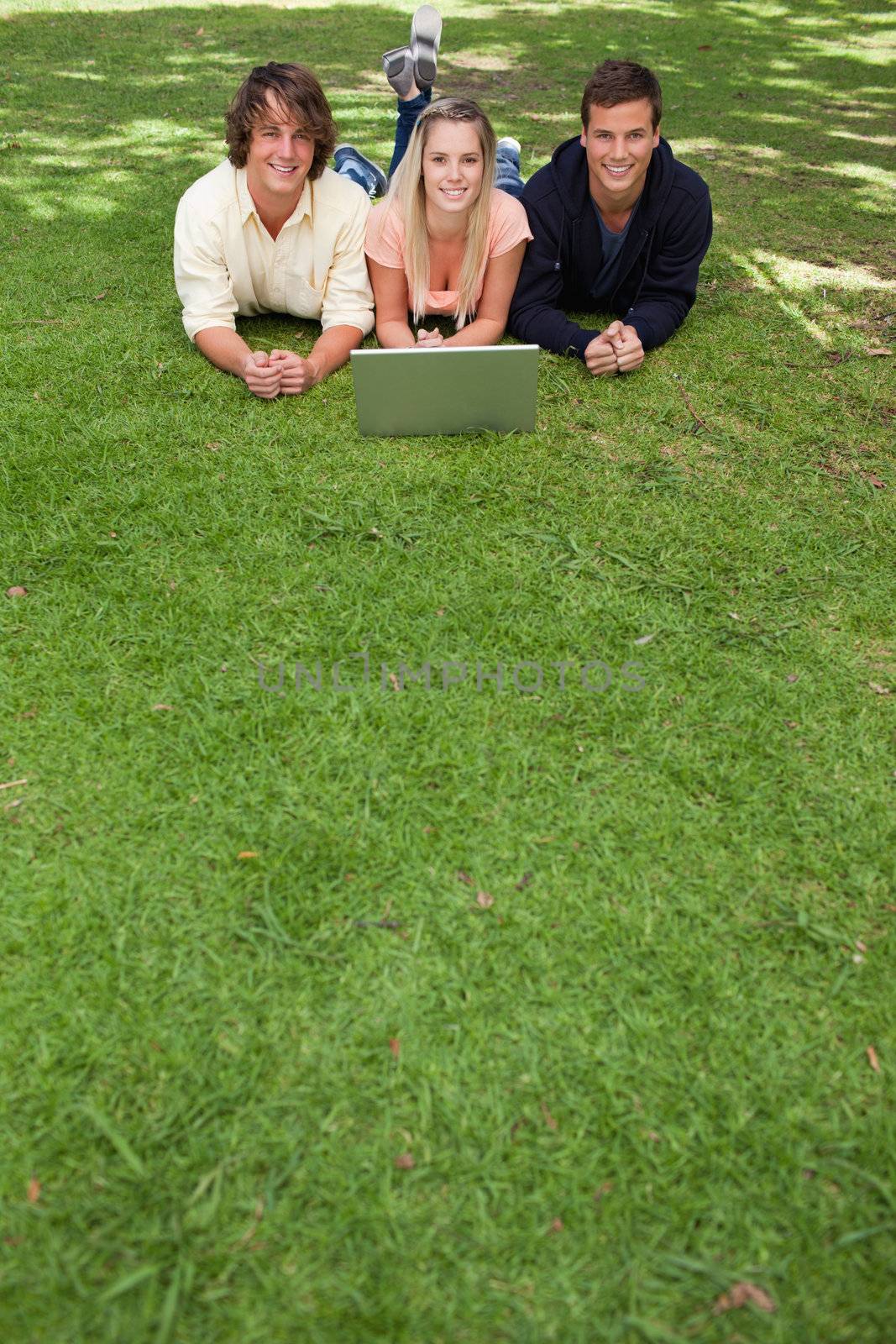 Three smiling students lying in a park with a laptop
