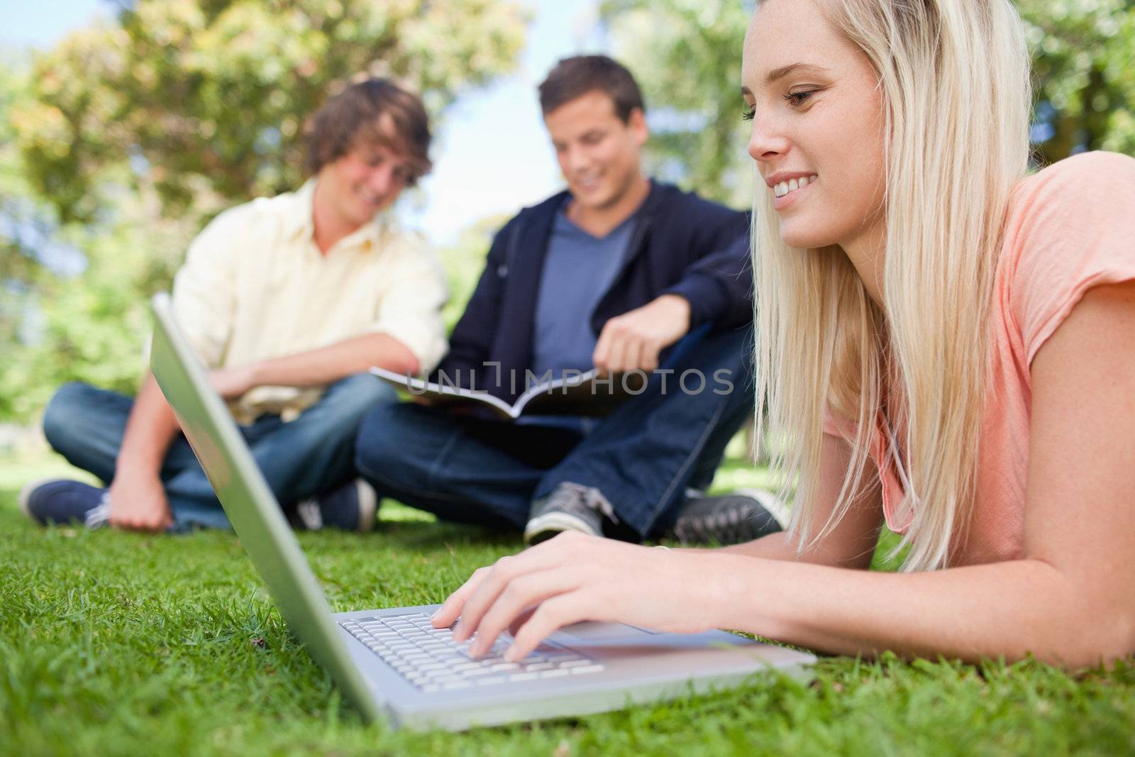 Girl using a laptop while lying in a park with friends in background
