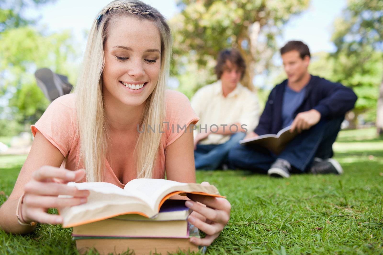 Girl lying while reading books in a park with friends in background