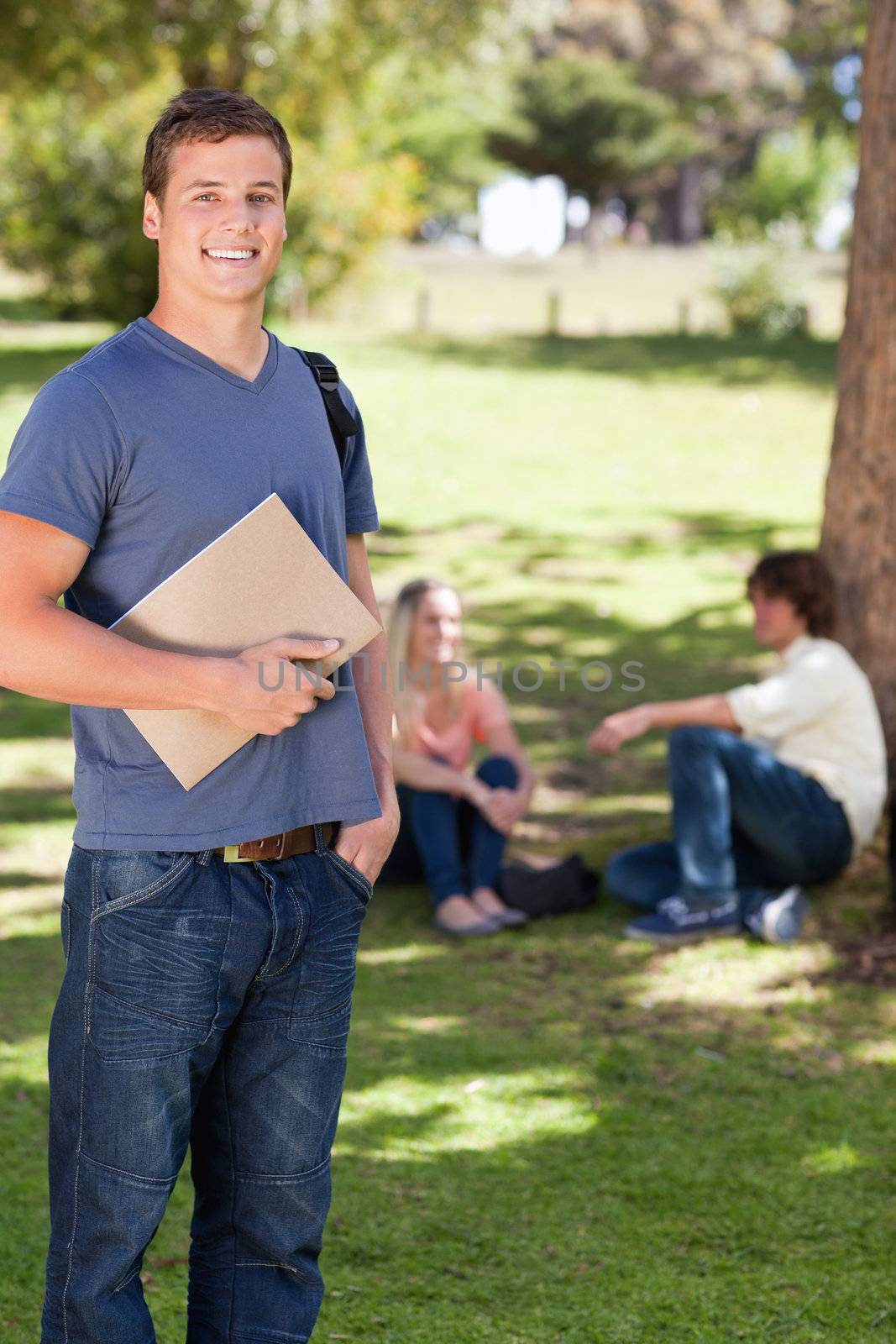 Student smiling while holding a textbook in a park with friends in background