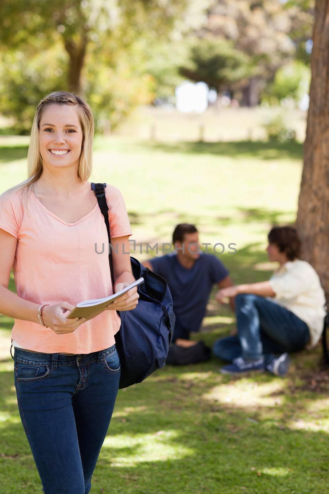 Portrait of a smiling female holding a textbook in a park with friends in background