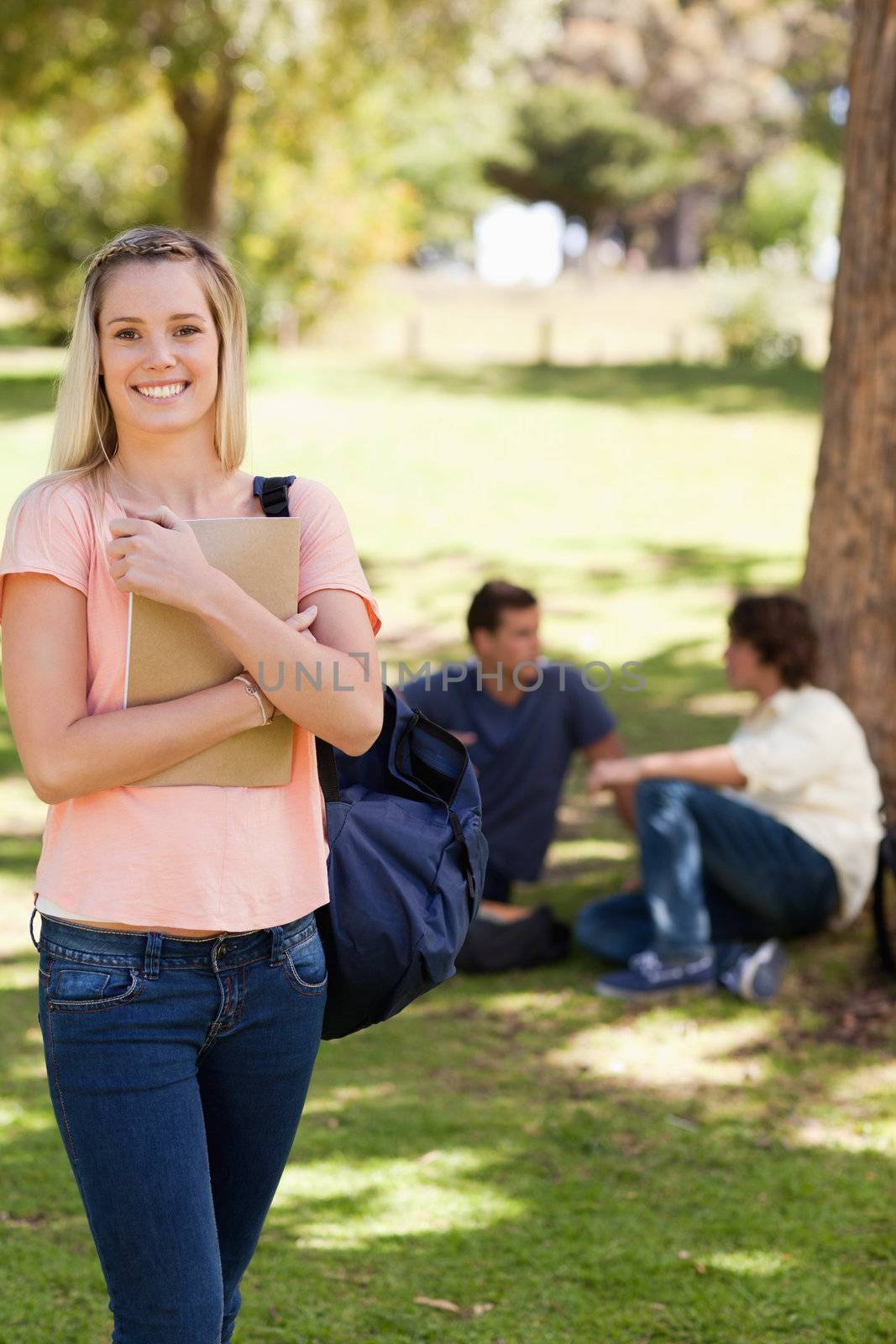 Smiling female holding a textbook in a park with friends in background