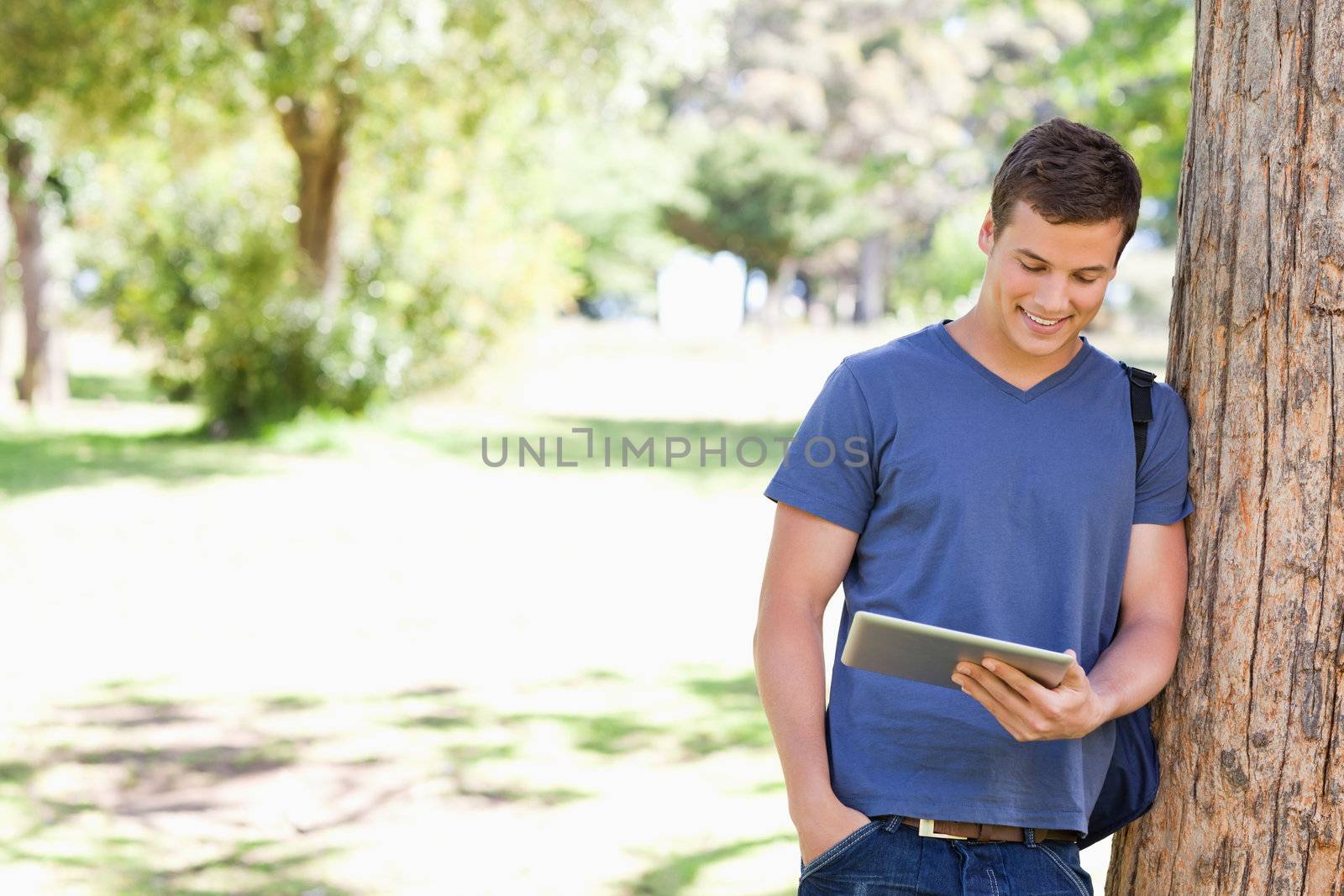 Student leaning against a tree with a touch pad in a park