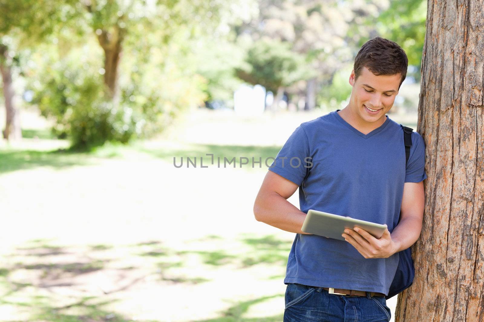 Student leaning against a tree while using a touch pad in a park