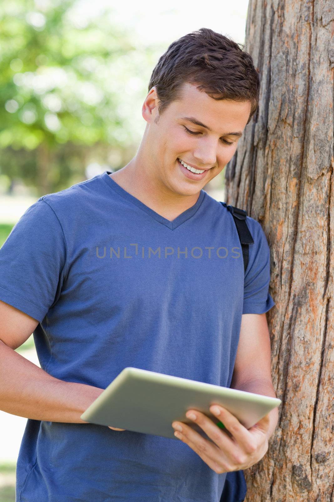Close-up of a young man leaning against a tree while using a touch pad in a park
