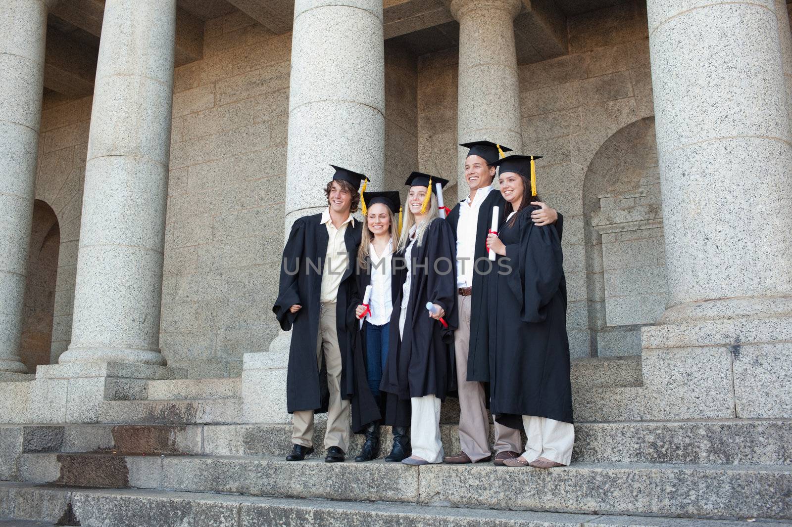 Five graduates posing while holding their diploma in front of the university