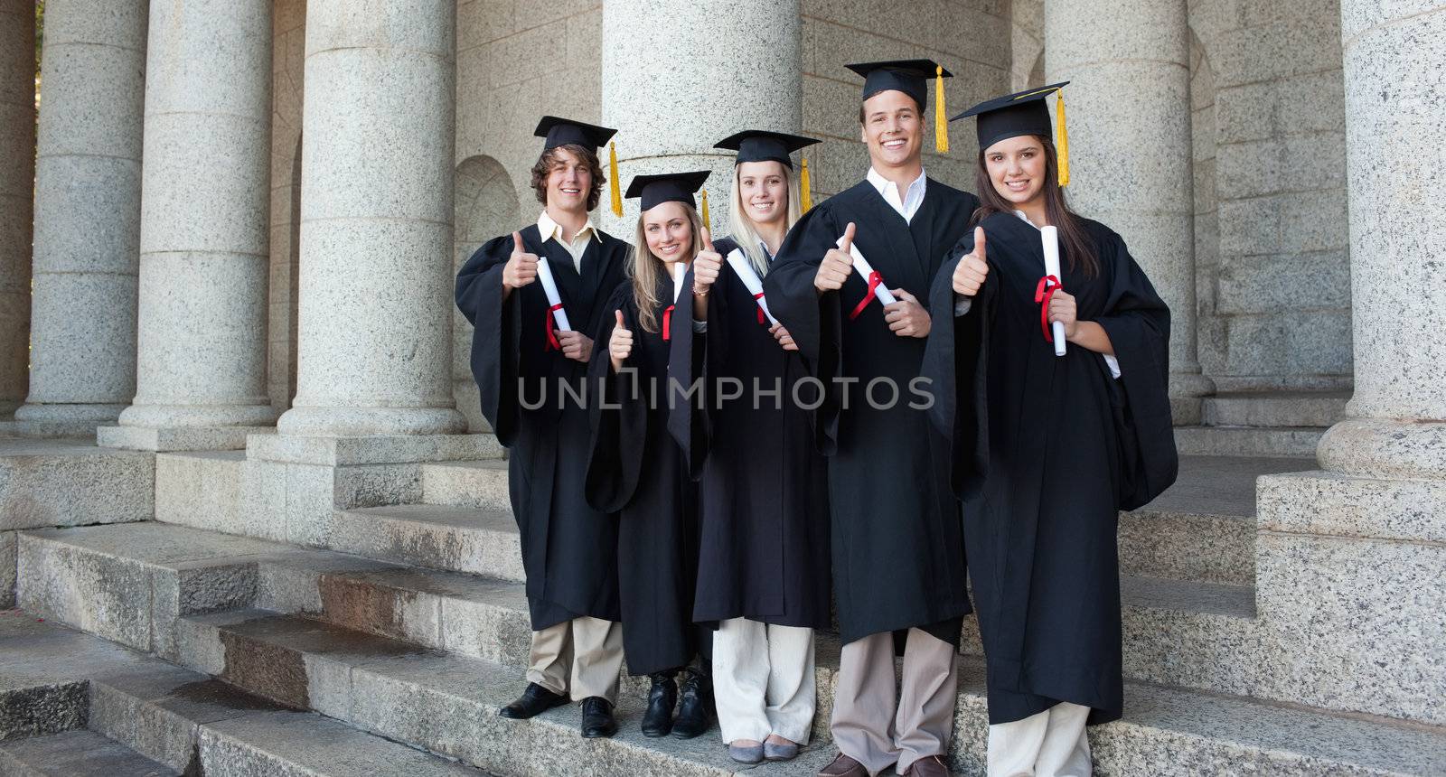 Five happy graduates posing the thumb-up in front of the university