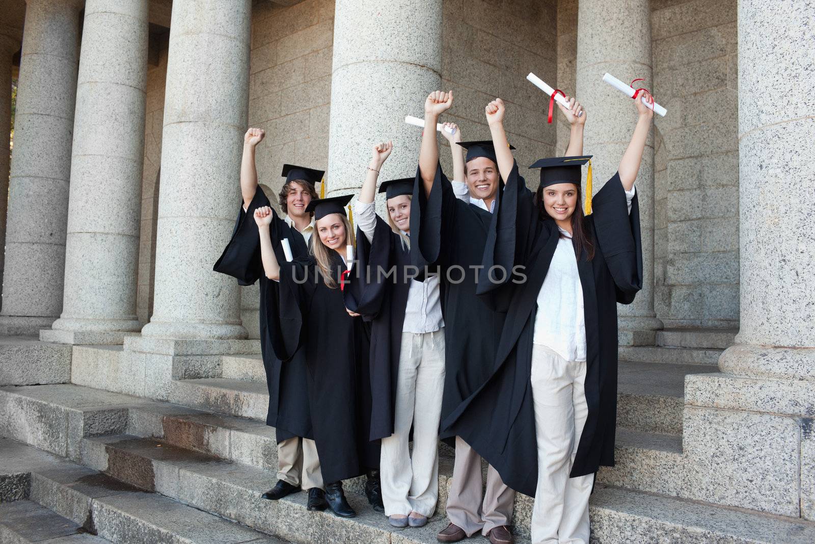Five happy graduates posing the arms raised in front of the university