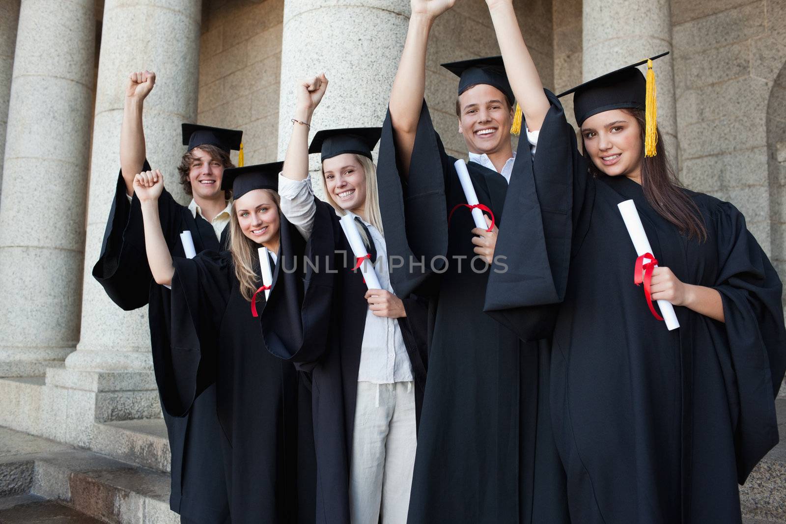 Five happy graduates posing the arm raised in front of the university