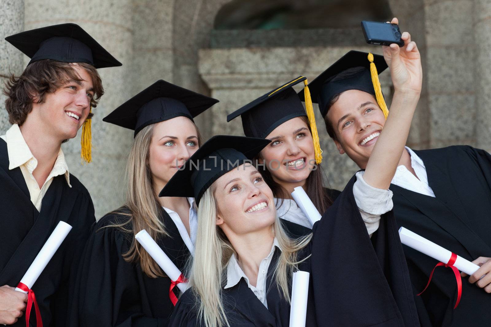 Close-up of five graduates taking a picture of themselves in front of the university
