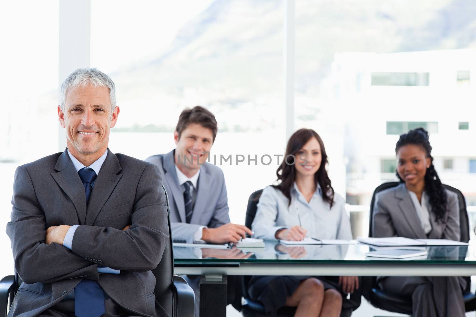 Mature smiling manager sitting with his arms crossed and with his team behind him