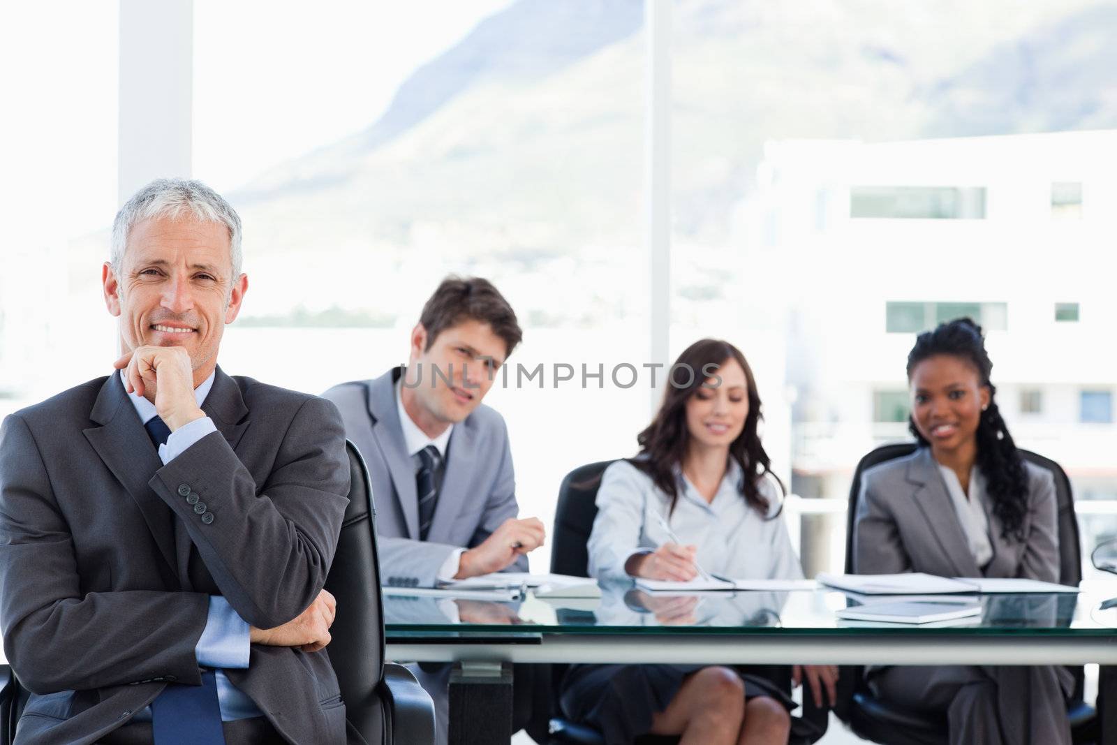 Mature smiling manager sitting with his hand on his chin while his team is looking at him