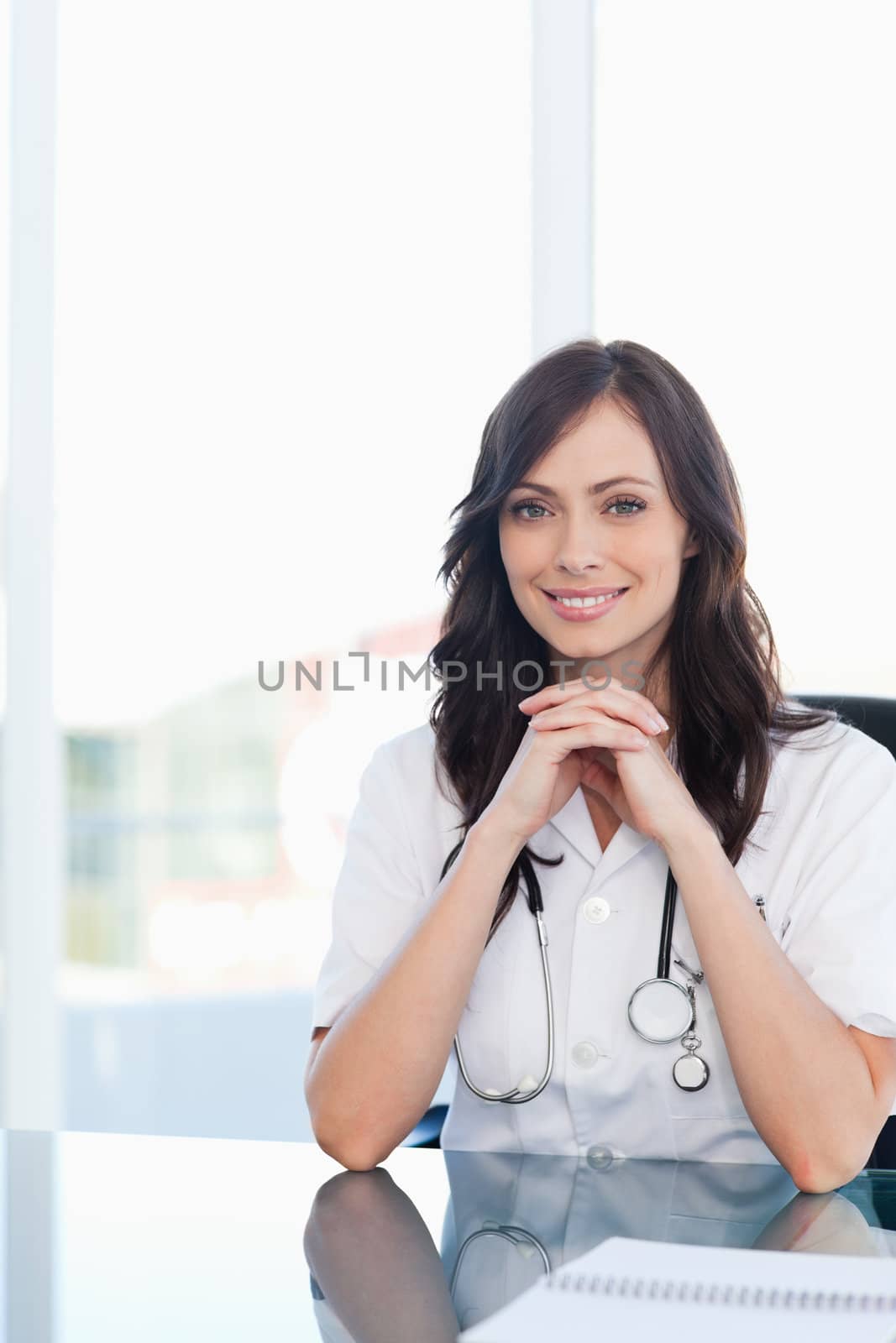 Smiling female doctor sitting at the desk with her hands crossed and her elbows resting on the table