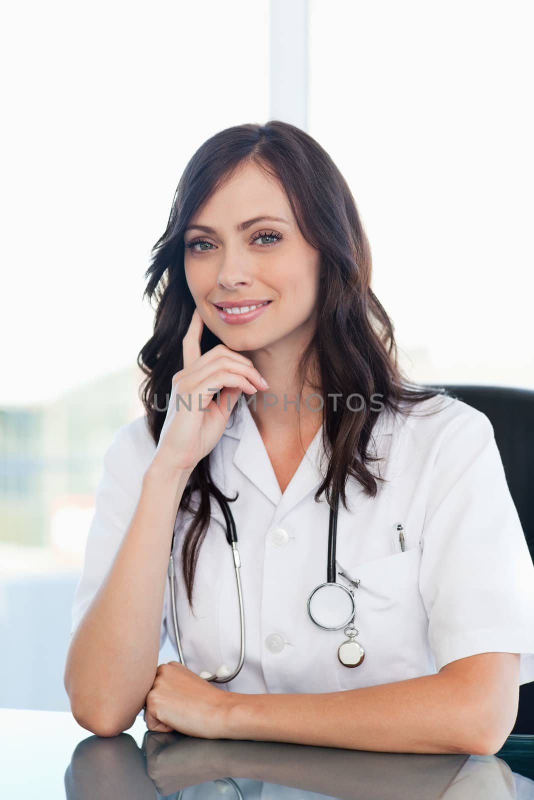 Smiling doctor sitting at the desk with her finger on her cheek by Wavebreakmedia