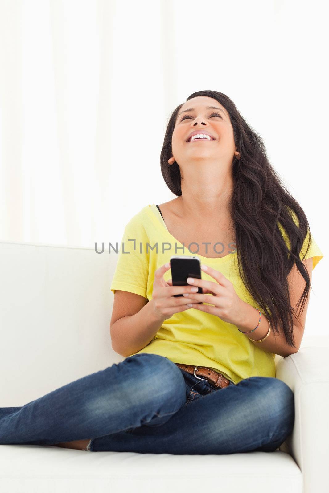 Happy Latino looking up while holding her smartphone being on a sofa