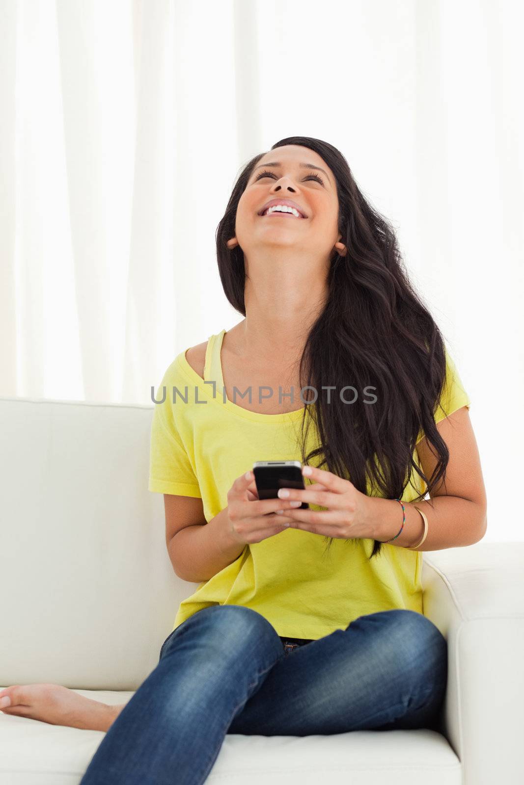 Happy Latino looking up while holding her cellphone being on a sofa