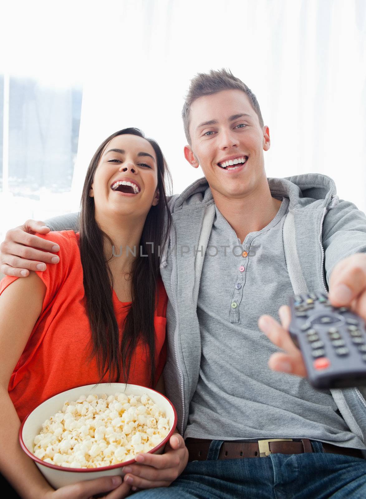 A couple sitting on the couch laughing as they have popcorn and look into the camera