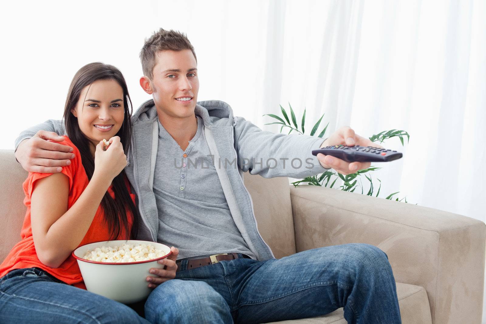 A smiling couple looking at the camera as they eat popcorn and use the tv remote