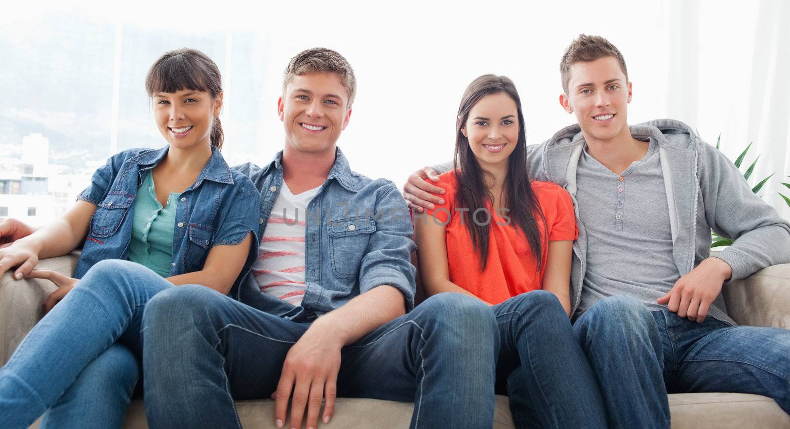 Two smiling couples sit on the couch beside one another as they look forward into the camera